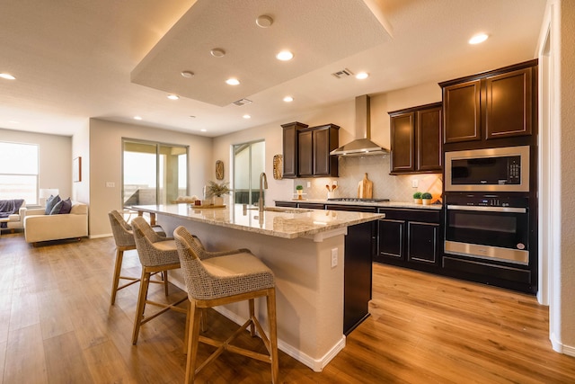 kitchen featuring stainless steel appliances, a sink, open floor plan, wall chimney exhaust hood, and a center island with sink