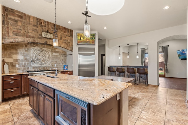 kitchen featuring built in appliances, light stone counters, decorative backsplash, a center island with sink, and decorative light fixtures