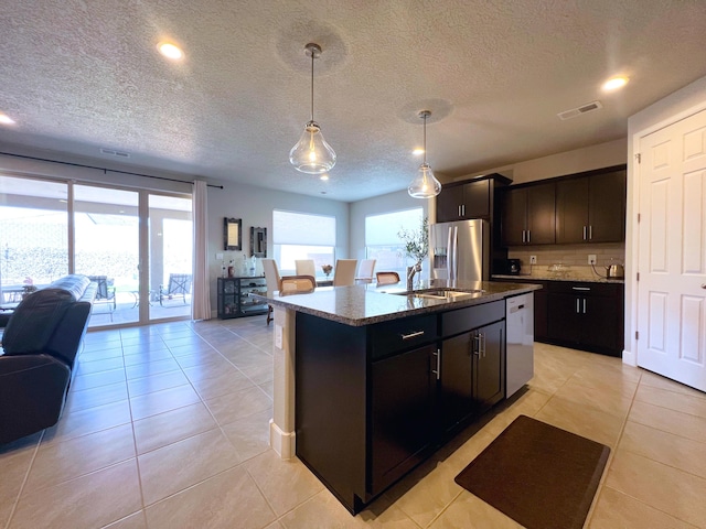 kitchen featuring stainless steel refrigerator with ice dispenser, decorative light fixtures, light tile patterned floors, an island with sink, and dark stone counters