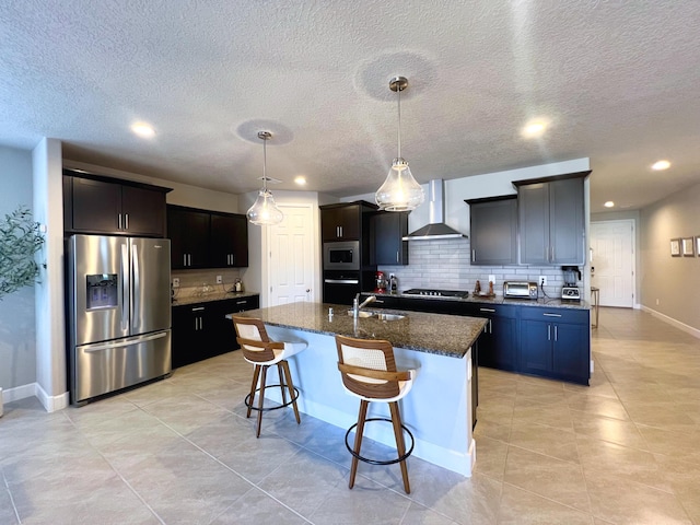 kitchen featuring tasteful backsplash, an island with sink, appliances with stainless steel finishes, and wall chimney range hood