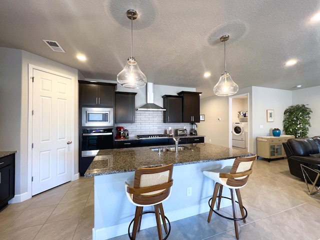 kitchen featuring sink, appliances with stainless steel finishes, an island with sink, washer / clothes dryer, and wall chimney range hood