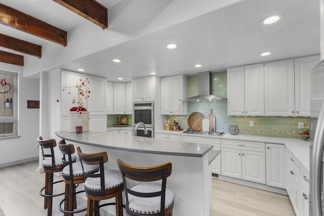 kitchen featuring wall chimney exhaust hood, white cabinetry, stainless steel appliances, beam ceiling, and backsplash