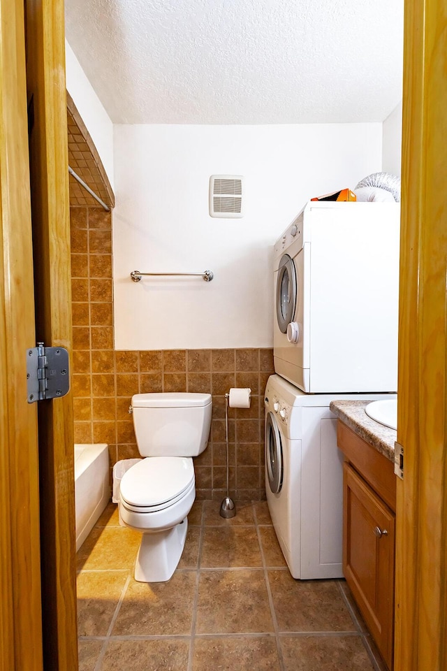 full bathroom with stacked washer and clothes dryer, toilet, a textured ceiling, tile walls, and vanity