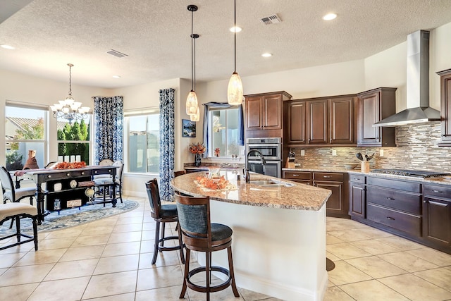 kitchen with stainless steel appliances, sink, an island with sink, and wall chimney range hood