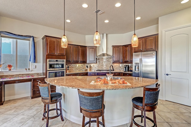 kitchen with a kitchen island with sink, light stone counters, stainless steel appliances, and wall chimney exhaust hood