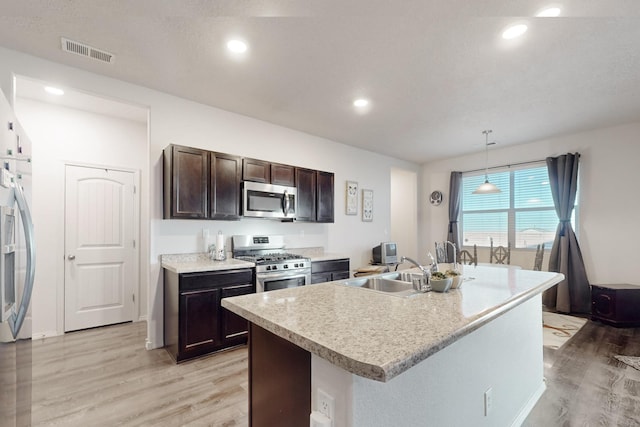 kitchen with appliances with stainless steel finishes, decorative light fixtures, sink, light hardwood / wood-style floors, and a center island with sink