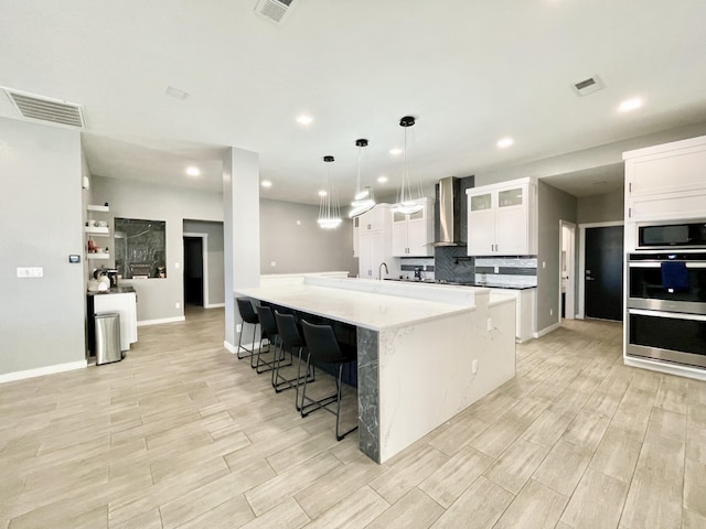 kitchen featuring a breakfast bar, white cabinets, hanging light fixtures, stainless steel double oven, and wall chimney exhaust hood