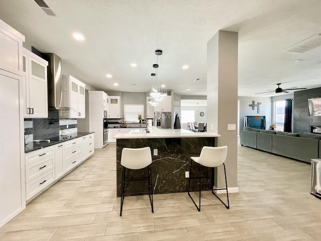 kitchen featuring stainless steel refrigerator with ice dispenser, wall chimney exhaust hood, a breakfast bar, a center island with sink, and white cabinets