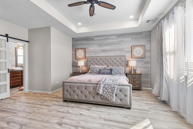 bedroom featuring ceiling fan, a tray ceiling, a textured ceiling, a barn door, and light wood-type flooring