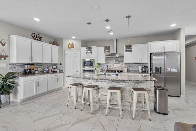 kitchen featuring appliances with stainless steel finishes, hanging light fixtures, a center island with sink, light stone countertops, and wall chimney range hood