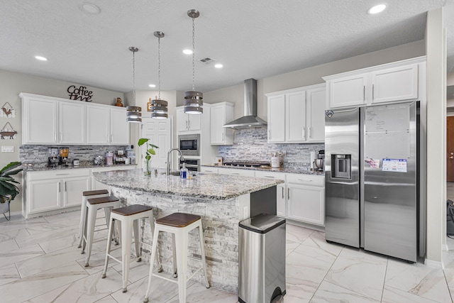 kitchen featuring pendant lighting, an island with sink, light stone counters, stainless steel appliances, and wall chimney exhaust hood