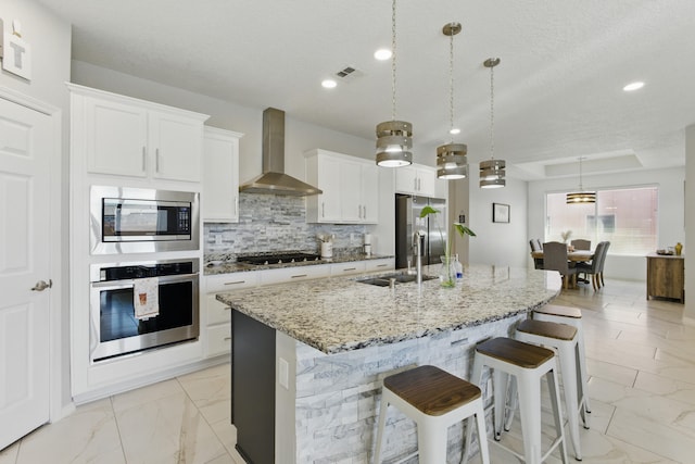 kitchen featuring an island with sink, a sink, stainless steel appliances, wall chimney exhaust hood, and marble finish floor