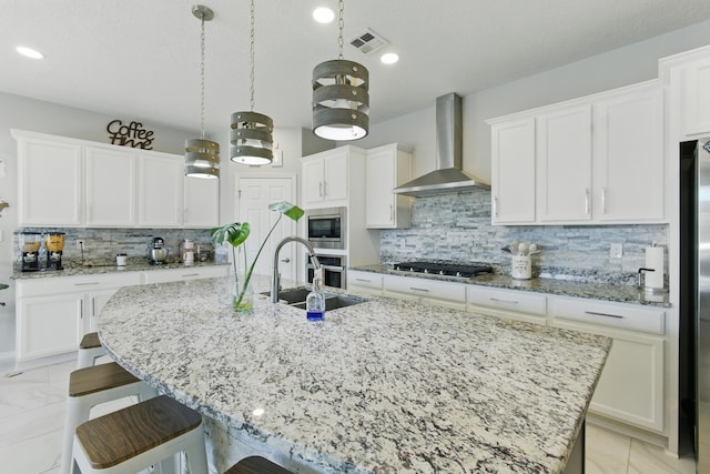 kitchen with visible vents, an island with sink, a sink, stainless steel appliances, and wall chimney range hood