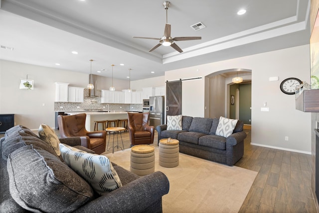 living room with ceiling fan, a barn door, light hardwood / wood-style floors, and a tray ceiling