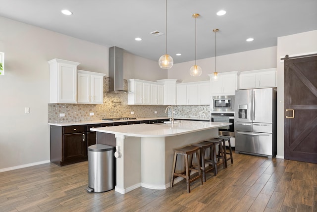 kitchen featuring white cabinetry, stainless steel appliances, a barn door, a center island with sink, and wall chimney exhaust hood