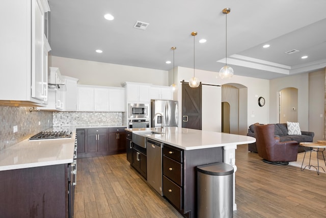 kitchen with tasteful backsplash, decorative light fixtures, an island with sink, stainless steel appliances, and white cabinets