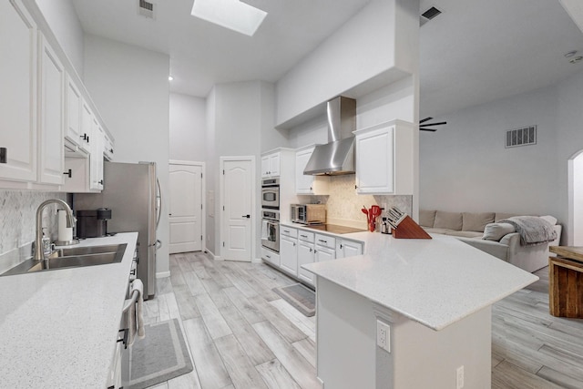 kitchen featuring wall chimney range hood, sink, a skylight, white cabinets, and kitchen peninsula