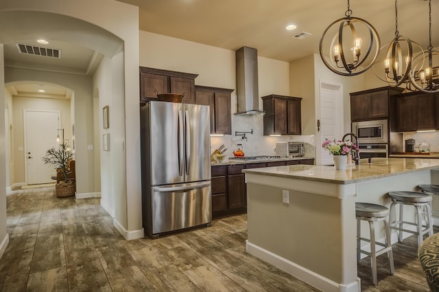 kitchen with appliances with stainless steel finishes, a kitchen bar, wall chimney range hood, light stone countertops, and a center island with sink