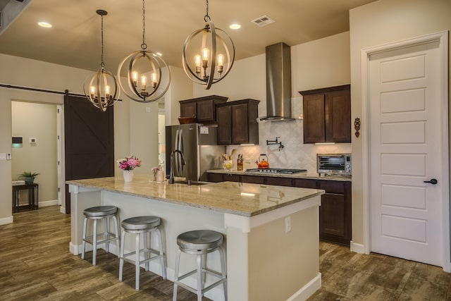 kitchen with decorative light fixtures, appliances with stainless steel finishes, an island with sink, a barn door, and wall chimney range hood