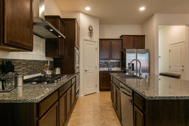 kitchen featuring a sink, dark brown cabinets, appliances with stainless steel finishes, wall chimney exhaust hood, and an island with sink