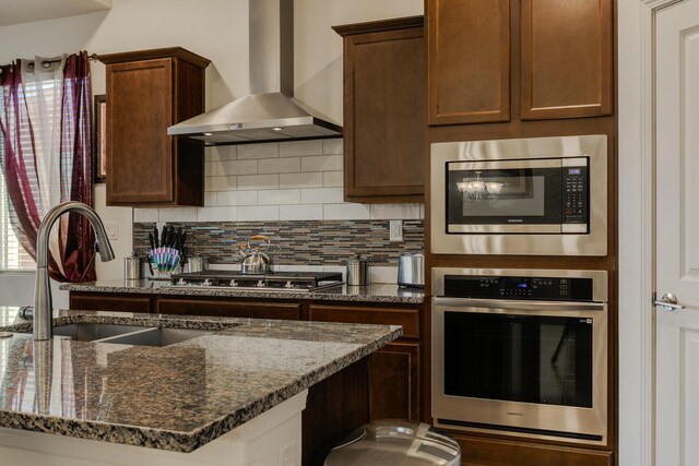 kitchen with appliances with stainless steel finishes, a sink, an island with sink, dark brown cabinets, and wall chimney exhaust hood