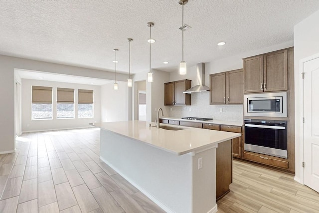 kitchen featuring appliances with stainless steel finishes, pendant lighting, sink, a kitchen island with sink, and wall chimney exhaust hood