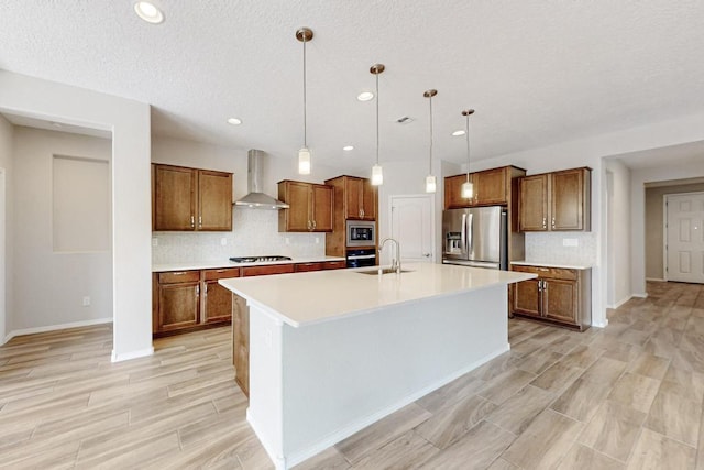 kitchen featuring wall chimney range hood, sink, a kitchen island with sink, hanging light fixtures, and stainless steel appliances