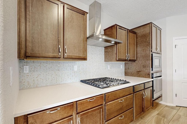 kitchen with a textured ceiling, appliances with stainless steel finishes, light hardwood / wood-style floors, decorative backsplash, and wall chimney range hood