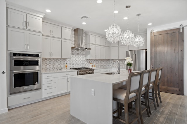 kitchen featuring pendant lighting, wall chimney range hood, sink, appliances with stainless steel finishes, and a kitchen island with sink