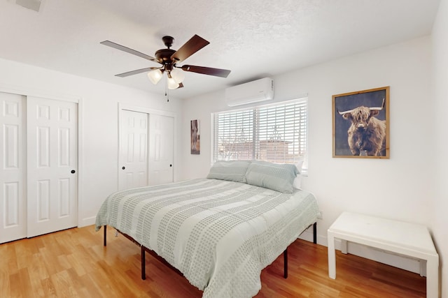 bedroom with a textured ceiling, an AC wall unit, two closets, ceiling fan, and hardwood / wood-style floors