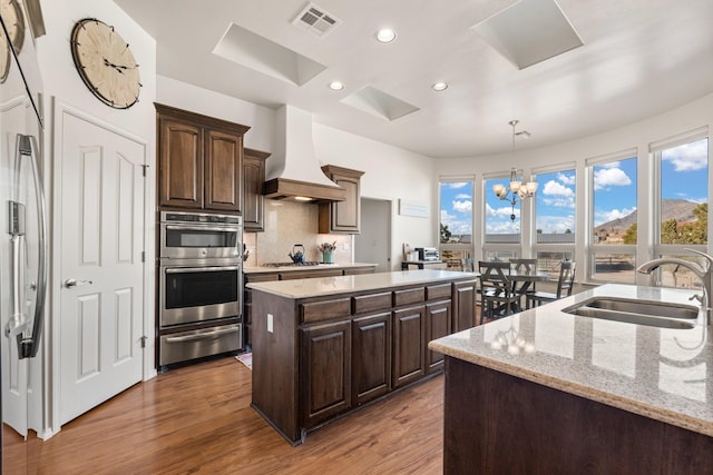 kitchen with sink, dark brown cabinets, a center island with sink, custom range hood, and stainless steel appliances