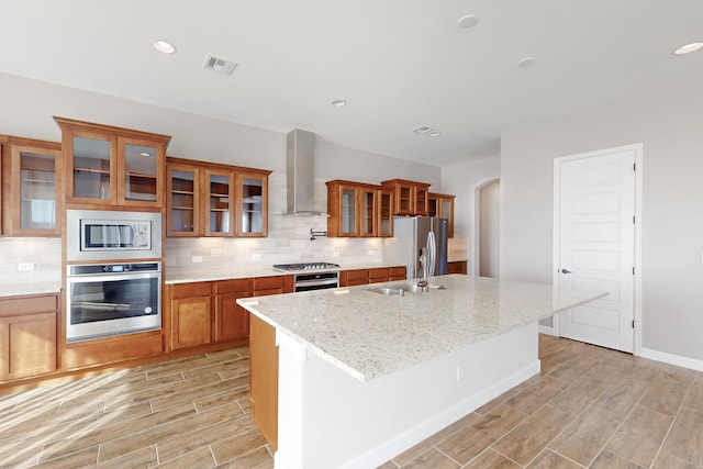 kitchen featuring wall chimney range hood, appliances with stainless steel finishes, backsplash, light stone counters, and a center island with sink