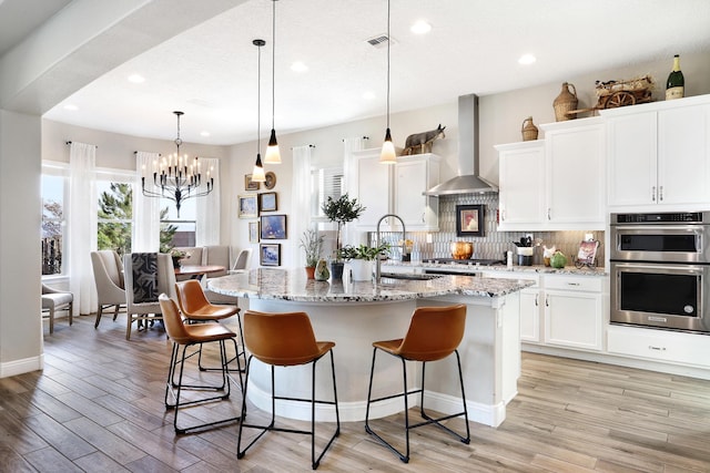 kitchen featuring pendant lighting, an island with sink, sink, white cabinets, and wall chimney exhaust hood