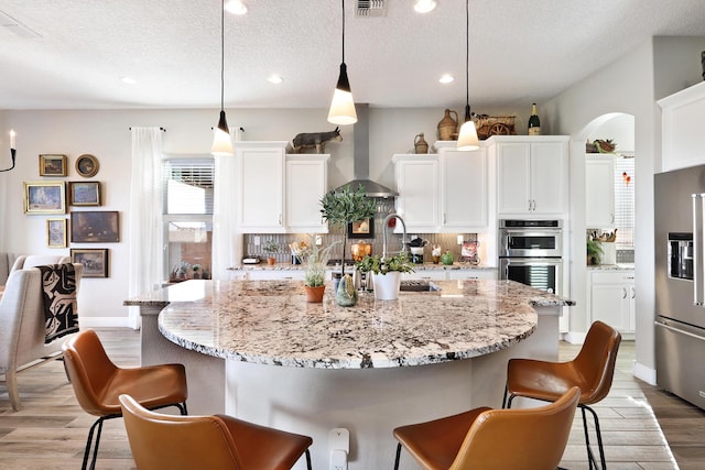 kitchen featuring wall chimney exhaust hood, hanging light fixtures, appliances with stainless steel finishes, a kitchen island with sink, and white cabinets