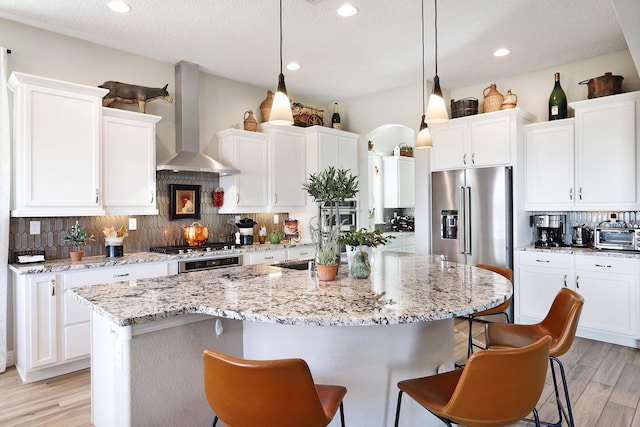 kitchen with pendant lighting, wall chimney exhaust hood, a kitchen island with sink, and white cabinets