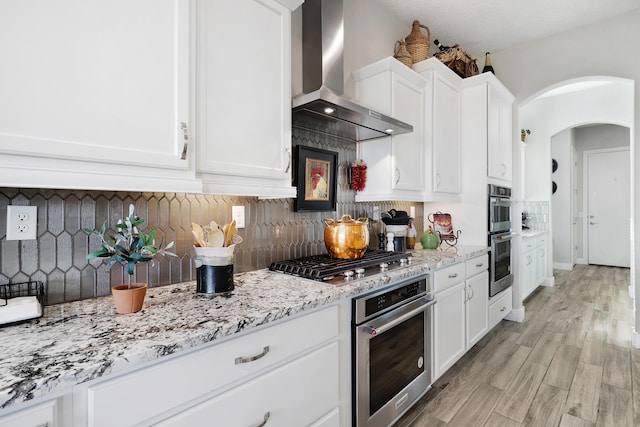 kitchen featuring wall chimney exhaust hood, white cabinets, stainless steel appliances, light stone countertops, and backsplash