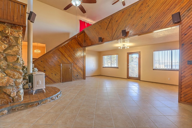 unfurnished living room featuring ceiling fan, a towering ceiling, wood walls, light tile patterned flooring, and a wood stove