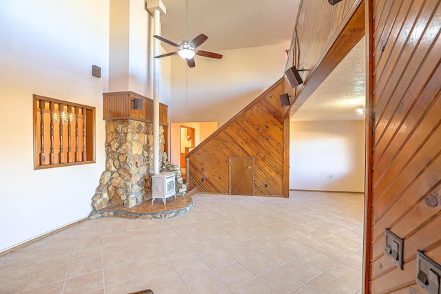 unfurnished living room with ceiling fan, wooden walls, a high ceiling, a textured ceiling, and a wood stove