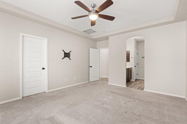 unfurnished bedroom with ceiling fan, light colored carpet, a tray ceiling, and ensuite bath