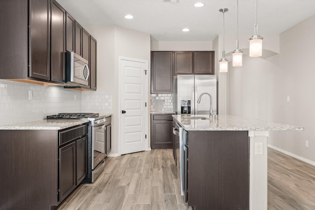 kitchen with appliances with stainless steel finishes, a kitchen island with sink, hanging light fixtures, dark brown cabinetry, and light wood-type flooring