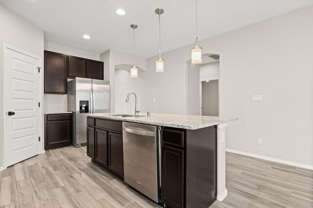 kitchen featuring sink, appliances with stainless steel finishes, an island with sink, decorative light fixtures, and light wood-type flooring