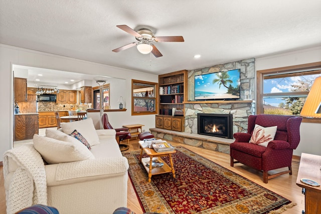 living area with a ceiling fan, light wood finished floors, ornamental molding, a stone fireplace, and a textured ceiling