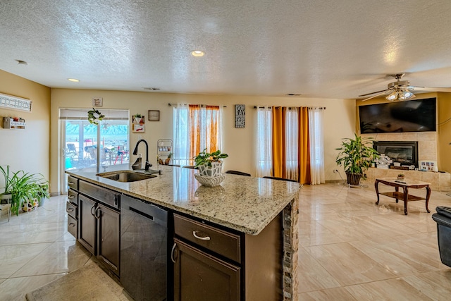 kitchen with a tile fireplace, dishwasher, sink, a kitchen island with sink, and light stone counters