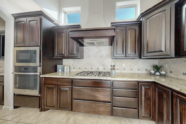 kitchen with light stone counters, dark brown cabinetry, stainless steel appliances, custom exhaust hood, and tasteful backsplash