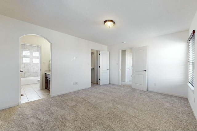 unfurnished bedroom featuring arched walkways, connected bathroom, light colored carpet, baseboards, and a walk in closet