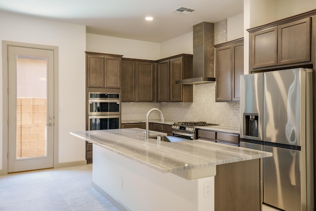 kitchen with appliances with stainless steel finishes, sink, light stone counters, a center island with sink, and wall chimney exhaust hood