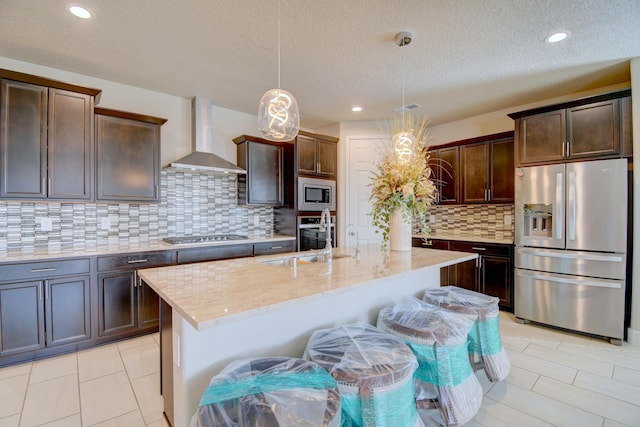 kitchen with a kitchen island with sink, hanging light fixtures, stainless steel appliances, dark brown cabinetry, and wall chimney exhaust hood