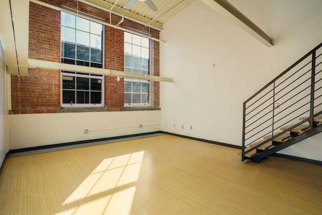 unfurnished living room featuring ceiling fan, hardwood / wood-style floors, beam ceiling, a high ceiling, and brick wall