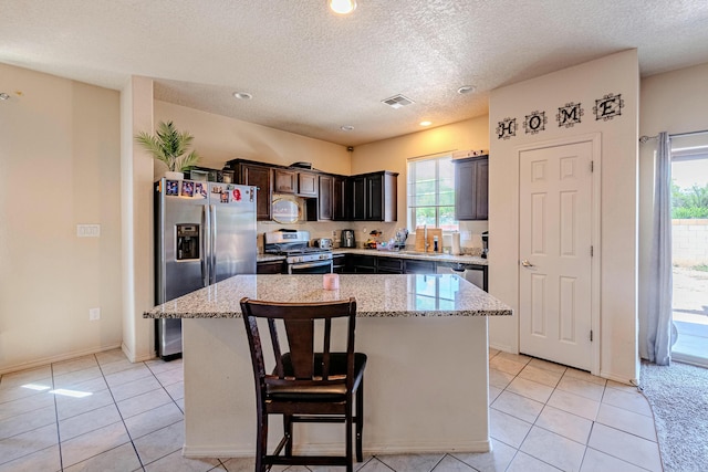 kitchen featuring dark brown cabinets, stainless steel appliances, a kitchen island, light stone countertops, and light tile patterned flooring