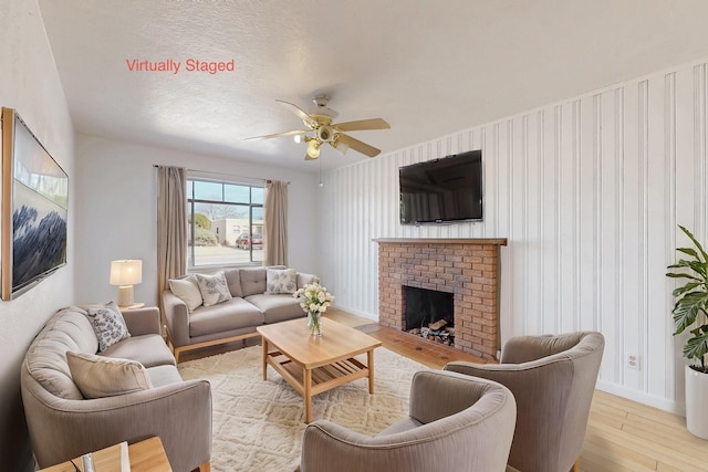 living room with ceiling fan, light hardwood / wood-style floors, a brick fireplace, and a textured ceiling